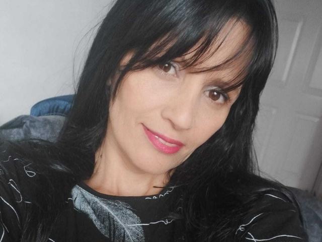 DulceHotShary with black hair, brown eyes, pink lipstick, smiling at the camera, wearing a black shirt with white lines, sitting in a blue chair.