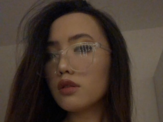 MishelBlessing has long dark hair, clear glasses, and full pink lips, looking serious with a slight pout. Her Asian features and light skin are visible.