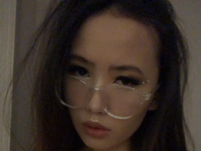 MishelBlessing with dark, messy hair, wearing transparent, oversized glasses, and slightly parted, nude-colored lips, looking serious in a dimly lit photo.