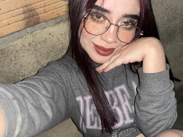 AdaraForest, avec des lunettes, du rouge à lèvres foncé, un anneau de nez et de longs cheveux bruns, portant un sweat à capuche gris "BRESCA", avec la tête appuyée sur sa main, souriant à l'appareil photo..