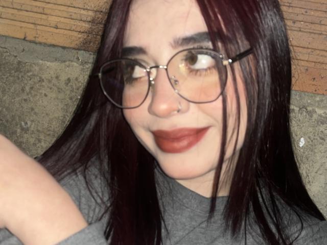 AdaraForest, avec ses cheveux roux foncés, ses lunettes, son anneau de nez et son rouge à lèvres rouge foncé, souriant légèrement, portant une chemise grise, avec un bras visible au premier plan..