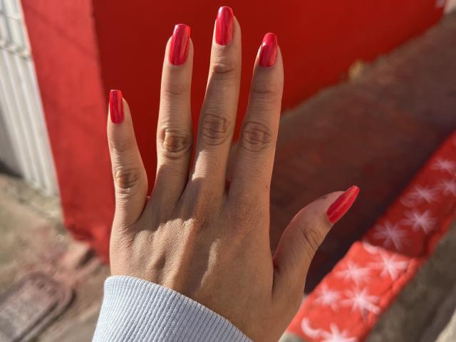 LillyTaylor's hand with shiny red nails, slightly curved fingers, and a light gray sleeve, against a bright red background. Sunlight highlights her nail polish.