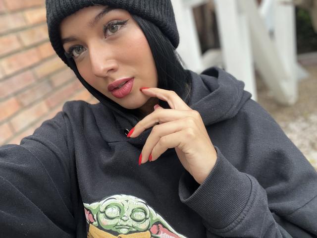 LillyTaylor with dark hair, wearing a black beanie and hoodie with a green Yoda graphic, red nails, and red lipstick, touching her chin with a slight smile.