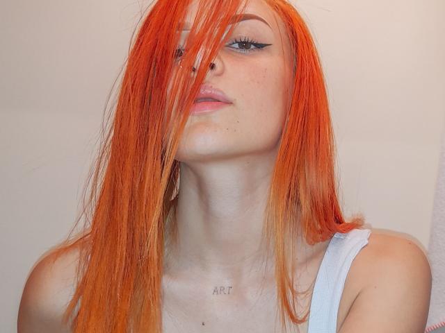CelesteBlazze has long orange hair covering one eye, wearing a white tank top, with black eyeliner and a nose ring, freckles on her face, and "ART" tattoo on her chest.