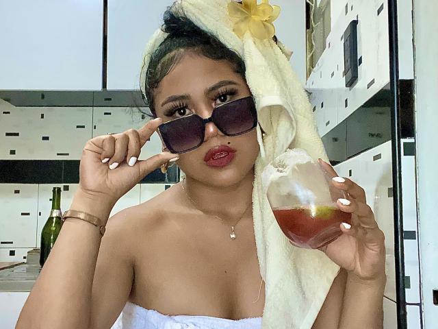 SophieSonyas with a towel on her head, dark sunglasses, red lipstick, holding a drink with a lemon slice, white towel, and yellow flower, showing off her bare tits.
