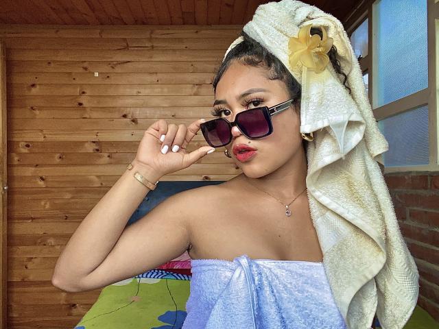 SophieSonyas with towel on head, yellow flower, dark sunglasses, red lips, white nails, blue towel, wooden ceiling, holding sunglasses, gold bracelet, necklace.