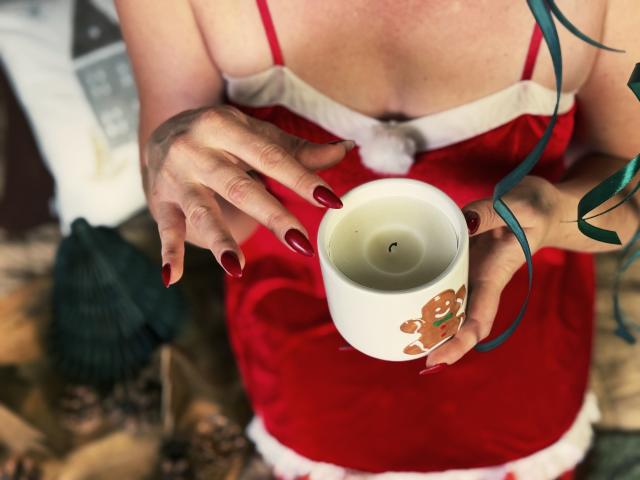 SnezhanaRells holds a white mug with a gingerbread cookie design, wearing a red Santa dress, showing her red-manicured nails and cleavage, with a green ribbon in her hair.
