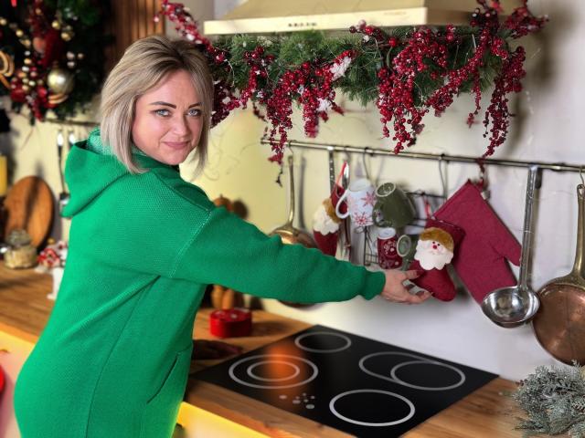 SnezhanaRells with blonde hair, green hoodie, reaching for a red Christmas stocking on a kitchen wall, holding a silver spoon, looking back with a slight smile.