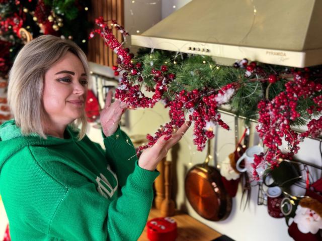 SnezhanaRells with blonde hair, green hoodie, holding red and green Christmas garland, smiling, hands up, kitchen, white stove, hanging pots, wooden floor, festive decor.