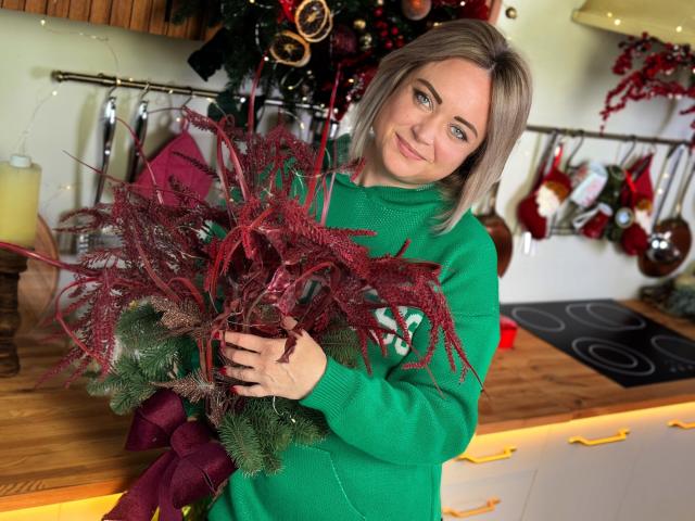 SnezhanaRells with blonde hair, green sweater, holding red and green Christmas wreath, smiling, standing in kitchen, wooden counter, yellow candle, Christmas decorations in background.