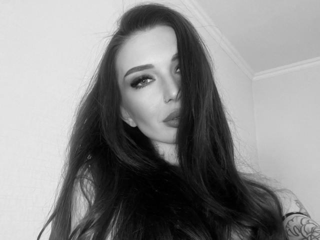 Black-and-white photo of sweetlessie-hot with long, messy dark hair, thick eyebrows, dramatic eyeliner, and full, pouty lips. Tattoo on right arm, slightly wavy hair covering nose.