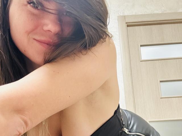 Ronete with messy brown hair, slight smile, exposed armpit, black strap top, leaning forward, showing small breasts and subtle cleavage, wooden door in background.
