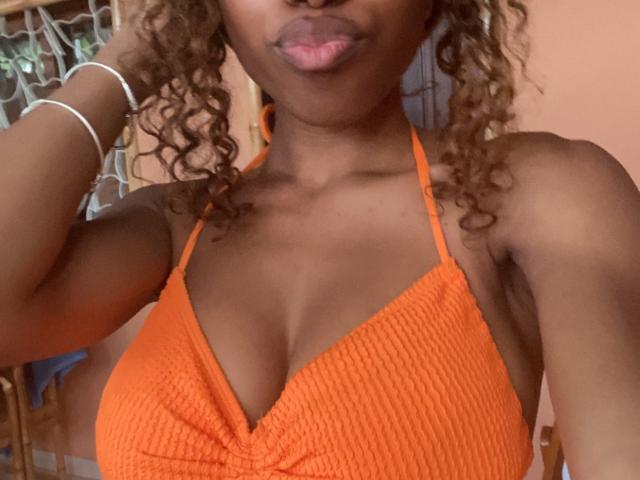 Ninah69 with curly brown hair, pouty pink lips, and dark skin, wearing an orange ribbed halter top that shows her large, cute boobs. She has a white bracelet.