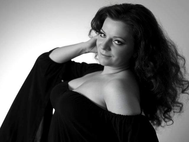 Delight-Alice with curly black hair, wearing an off-shoulder black dress, has her hand behind her head, showing her tits and a subtle smile in a black-and-white photo.