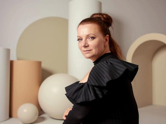 Delight-Alice with red hair in a bun, wearing a black top with ruffled shoulders, looking over her shoulder, standing in front of abstract geometric shapes.