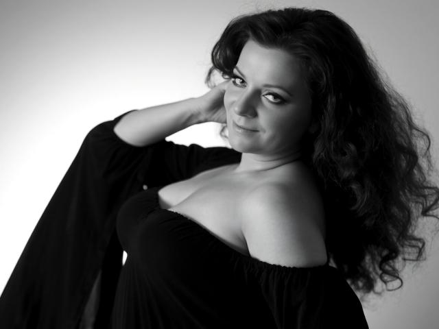 AliceXPerfect with wavy dark hair, black off-shoulder dress, and raised hand, smiling slightly, showcasing her boobs and soft cleavage in a grayscale, high-contrast photo.
