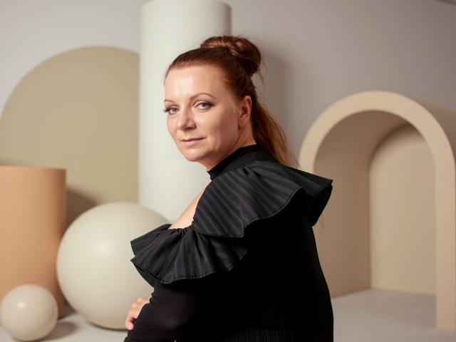 AliceXPerfect with red hair in a bun, wearing a black ruffled top, smiling at the camera, showing her tits slightly, in a modern, minimalist setting with geometric shapes.