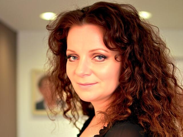 AliceXPerfect has curly brown hair, blue eyes, and a slight smile, wearing a black top, with soft, natural makeup and a warm, confident look.