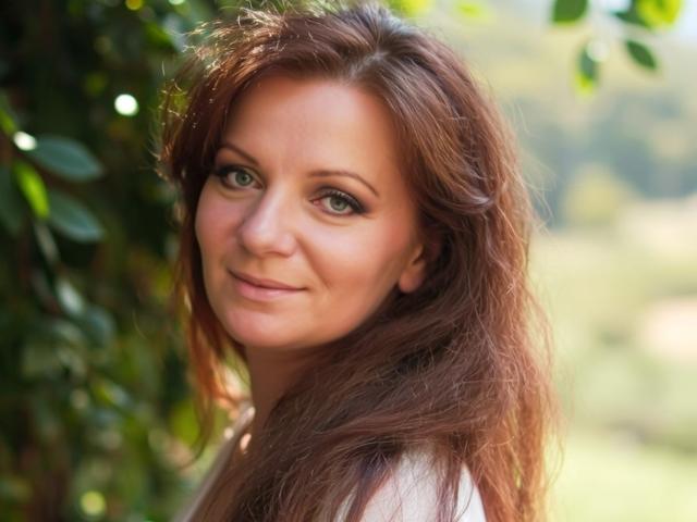AliceXPerfect has long, wavy brown hair and green eyes, smiling softly with a slight blush on her fair skin, looking directly at the camera in a sunny outdoor setting.