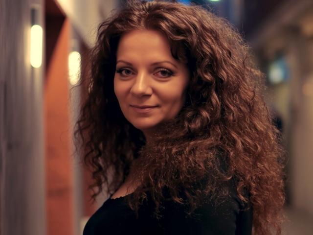 AliceXPerfect has curly brown hair, a slight smile, and is wearing a black top. She's standing in a dimly lit hallway with a warm, blurred background.