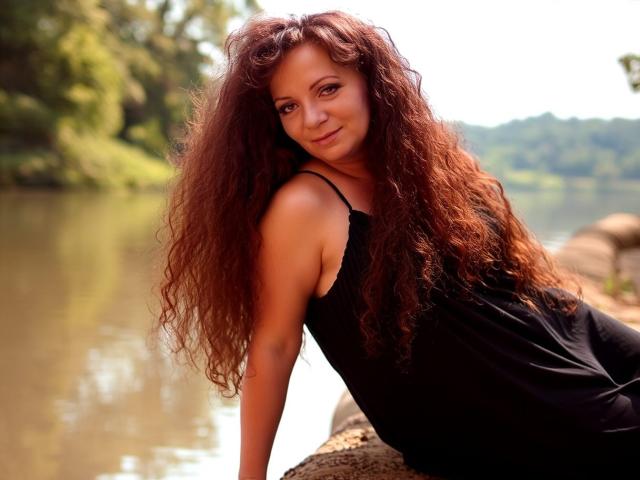 AliceXPerfect with long, curly brown hair, wearing a black dress, leaning on a rock by a river, smiling softly, showing her ass and cute face.