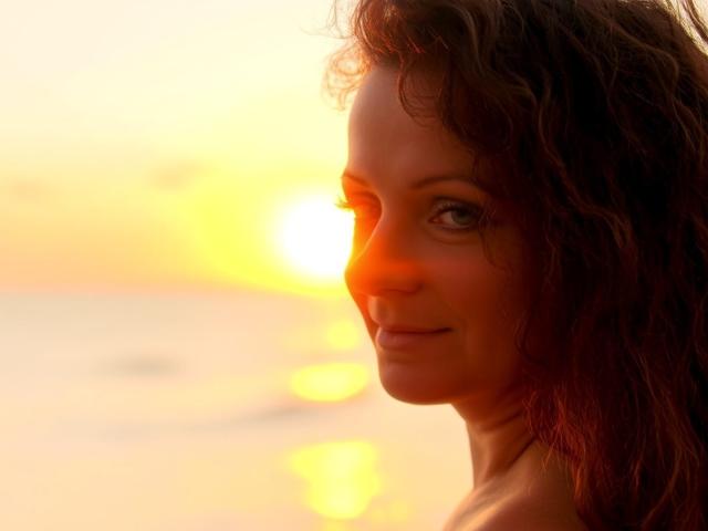 AliceXPerfect with curly brown hair, glowing orange sunset, and gentle smile, her ass slightly visible, looking over her shoulder, warm light highlighting her face and upper body.