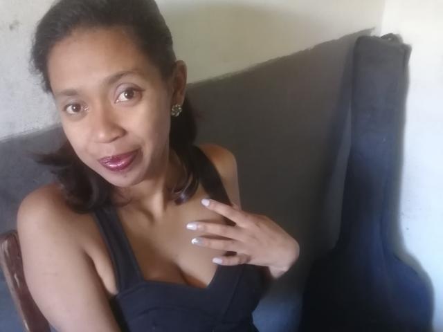 PetaKaily with brown skin and small tits, wearing a black dress, sitting on a grey chair, hand on chest, dark brown hair, shiny pink lipstick, and star earrings.