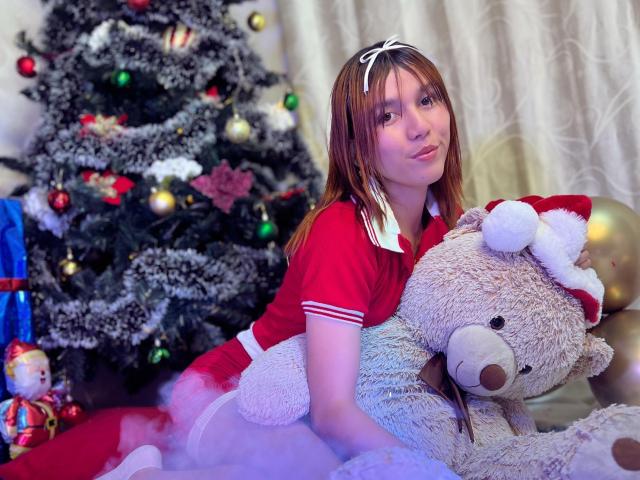 RachelJonier with brown hair, wearing a red Santa dress, white headband, and holding a large stuffed teddy bear with a Santa hat, sitting near a decorated Christmas tree.