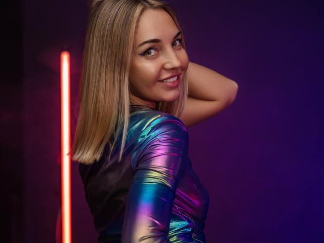 LettieSweet with blonde hair, smiling, looking back over her shoulder, wearing a shiny, metallic blue and purple top, hand in hair, neon red light on the left.