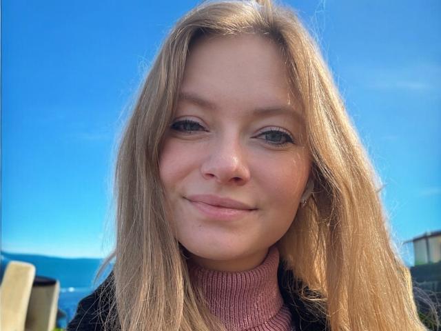 MillieLust has blonde hair, blue eyes, and a slight smile with a pink turtleneck. Her ass isn't visible, and her face is centered in the shot.