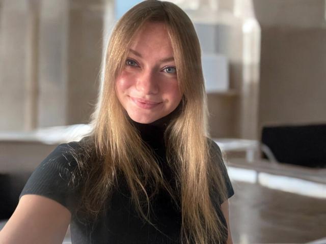 MillieLust with long blonde hair, black turtleneck, and a cute smile, standing in a sunlit room, showing her tits slightly in the soft light.