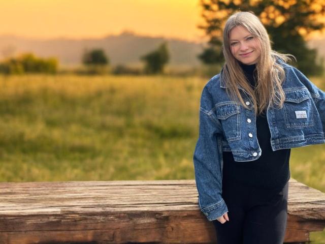 MillieLust with long blonde hair, wearing a denim jacket and black top, leaning on a wooden fence, smiling at sunset, grassy field in background.