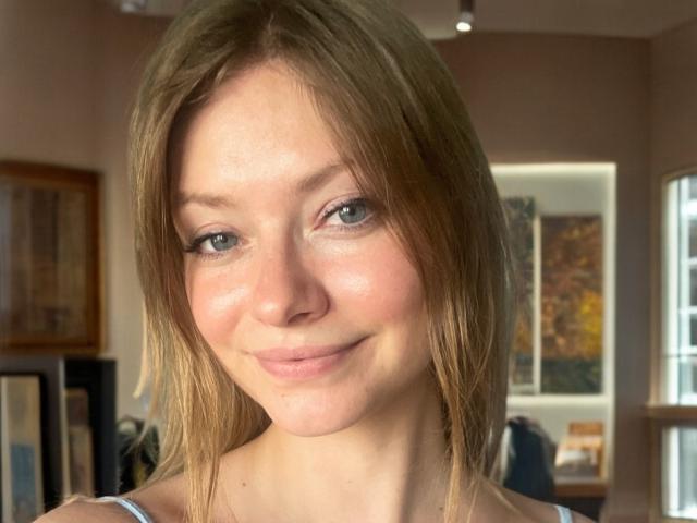 MillieLust has light brown hair, green eyes, and a slight smile with pinkish cheeks. She's wearing a white top, and her ass is not visible.