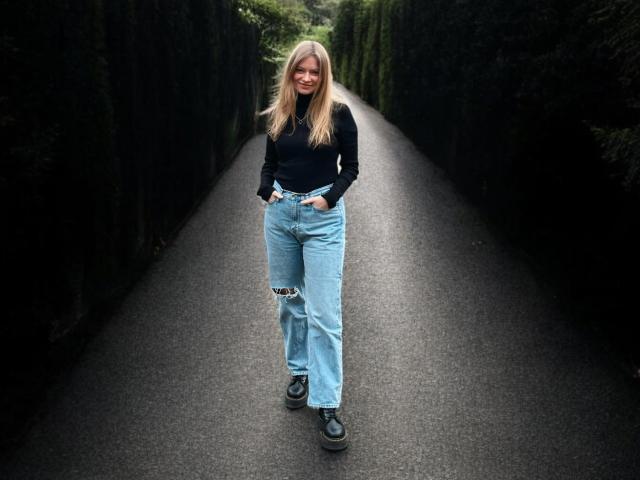 MillieLust stands on a dark, narrow road wearing a black long-sleeve shirt, ripped light blue jeans, and black shoes, smiling with hands in pockets.