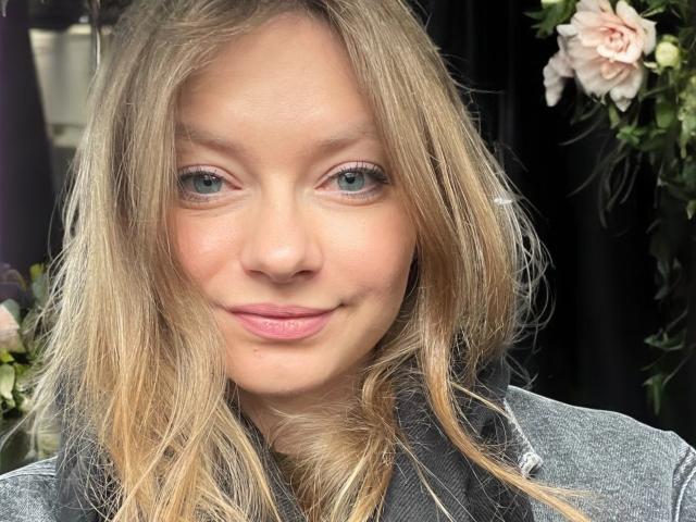 MillieLust has blonde hair, green eyes, and pink lips, wearing a grey jacket, with a subtle smile, and soft, natural makeup, looking slightly to the left.