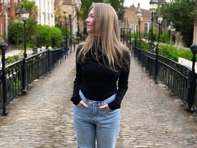 MillieLust has blonde hair, a black ribbed turtleneck, and blue jeans with hands in pockets, standing on a cobblestone bridge with black iron railings and vintage street lamps.