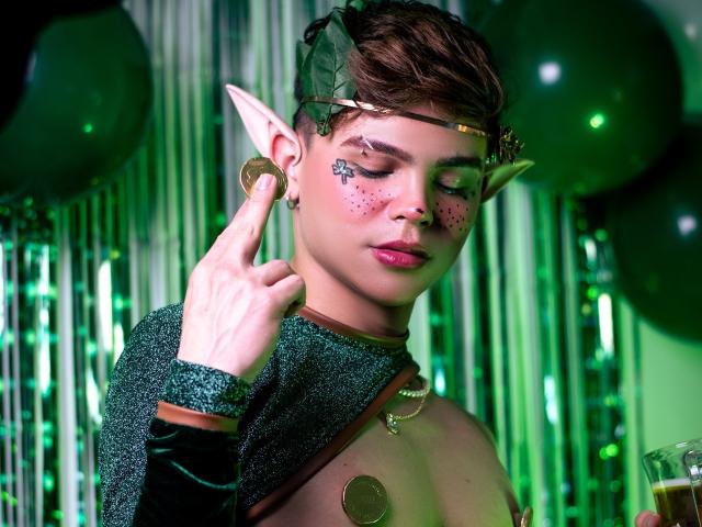KenniHotGuy with elf ears, green headband, glittery shirt, and coins on nipples, holding a drink, making a peace sign, with green and black balloons in the background.
