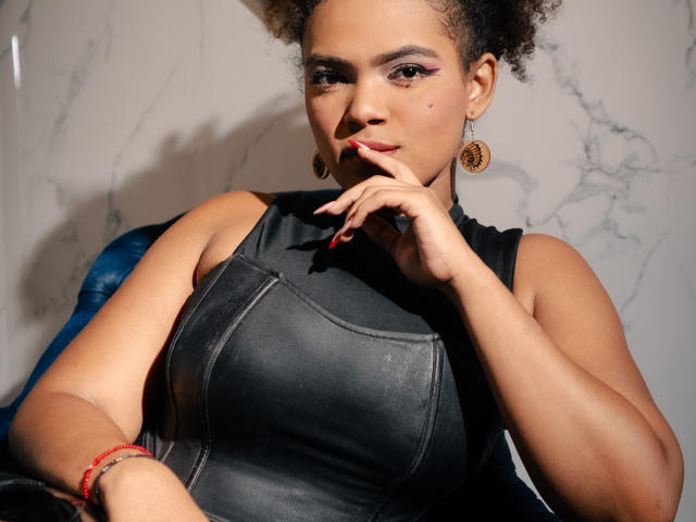 AliceJelanni with curly brown hair, wearing a black leather dress, touching her finger to her lips, earrings, and a red bracelet, looking confident and sexy.