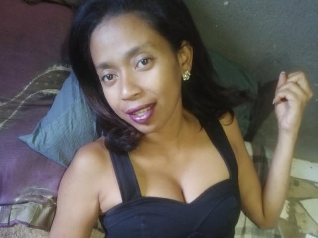PetaKaily with dark hair, brown skin, and small earrings, wearing a black top with visible tits, smiling with pink lipstick, right hand raised. Bed with purple and green pillows.