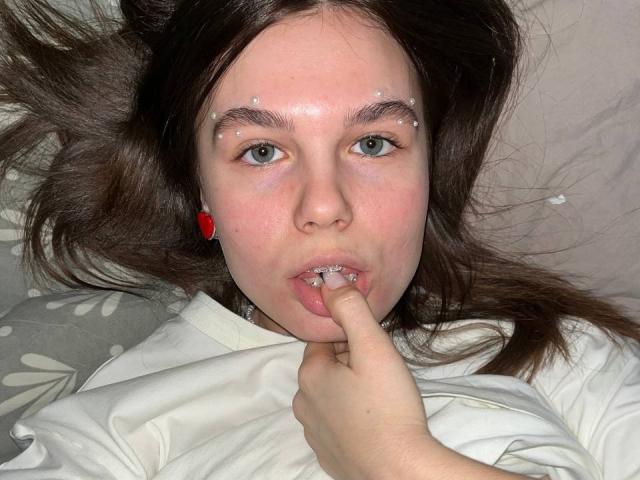LinaStones has light blue eyes, brown hair, and small studs in her eyebrows. She's biting her finger, with a red earring visible, wearing a white shirt.
