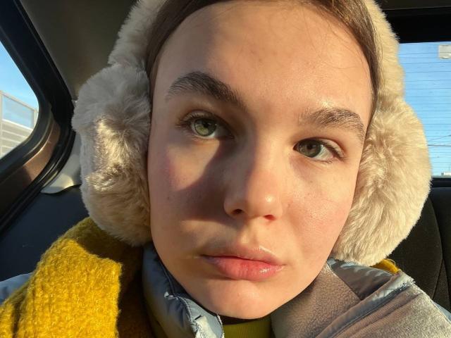 LinaStones has light green eyes, a cute face, and is wearing a fluffy white earmuff with a yellow vest. Sunlight highlights her fair skin and pink lips.