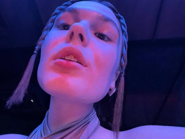 LinaStones has braided hair, glowing pink-blue skin, and shiny lips. She's wearing a halter top, looking up with a slightly open mouth. Low angle shot.