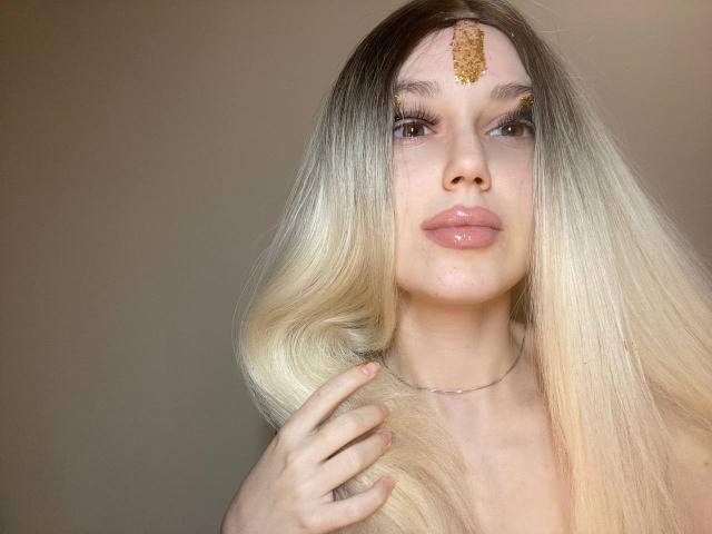 DitaVira with blonde ombre hair, gold forehead ornament, shiny pink lips, holding her hair, subtle necklace, soft brown eyes, looking at camera, slight smile.