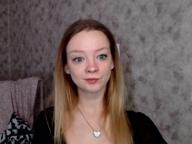 RadiantsLoves has blue eyes, long straight blonde hair, fair skin, and small tits. She wears a black top and a heart-shaped necklace. She's looking at the camera with a slight smile.