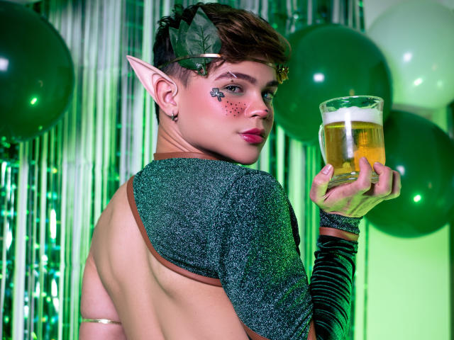 KenniHotGuy with elf ears, short brown hair, glittery green top, exposed back, holding beer, green face glitter, green balloons, and green striped arm, looking back.