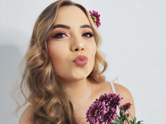ZoeTobon69 with pouty lips, blonde curly hair, pink eyeshadow, purple flower in hair, holding purple flowers, white strap top, cute and sexy, soft makeup.