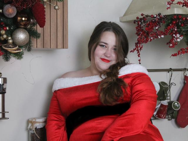 DaniellaGraceZZ, chubby girl with red lipstick, long brown hair, wearing off-shoulder red velvet Christmas dress with white fur, sitting, smiling, arms crossed, looking cute.
