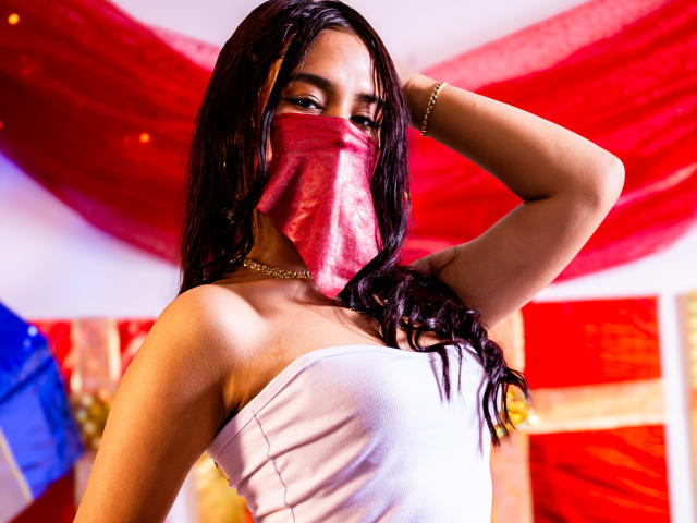 EmyXCute with dark hair, red mask, white strapless top, gold bracelet, and necklace, hand in hair, looking at camera, red and blue background, sexy, confident, curvy.