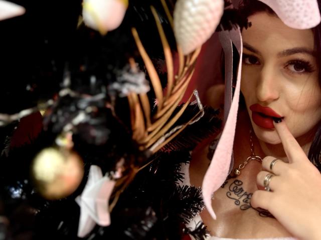 ArianaTattoos with red lips, pink hat, and finger in mouth, tattoo on chest, touching black Christmas tree with gold ornaments. Boobs partially visible, ring on finger.