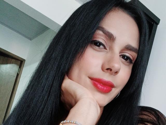 AnaisHilton has long black hair, brown eyes, red lipstick, and a cute smile. She's resting her chin on her hand with a subtle smirk. Her nose is small and cute.